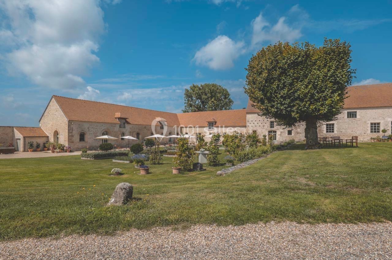 Bâtisse en pierre avec toit en tuiles, entourée d'un jardin verdoyant et d'une terrasse avec parasols.