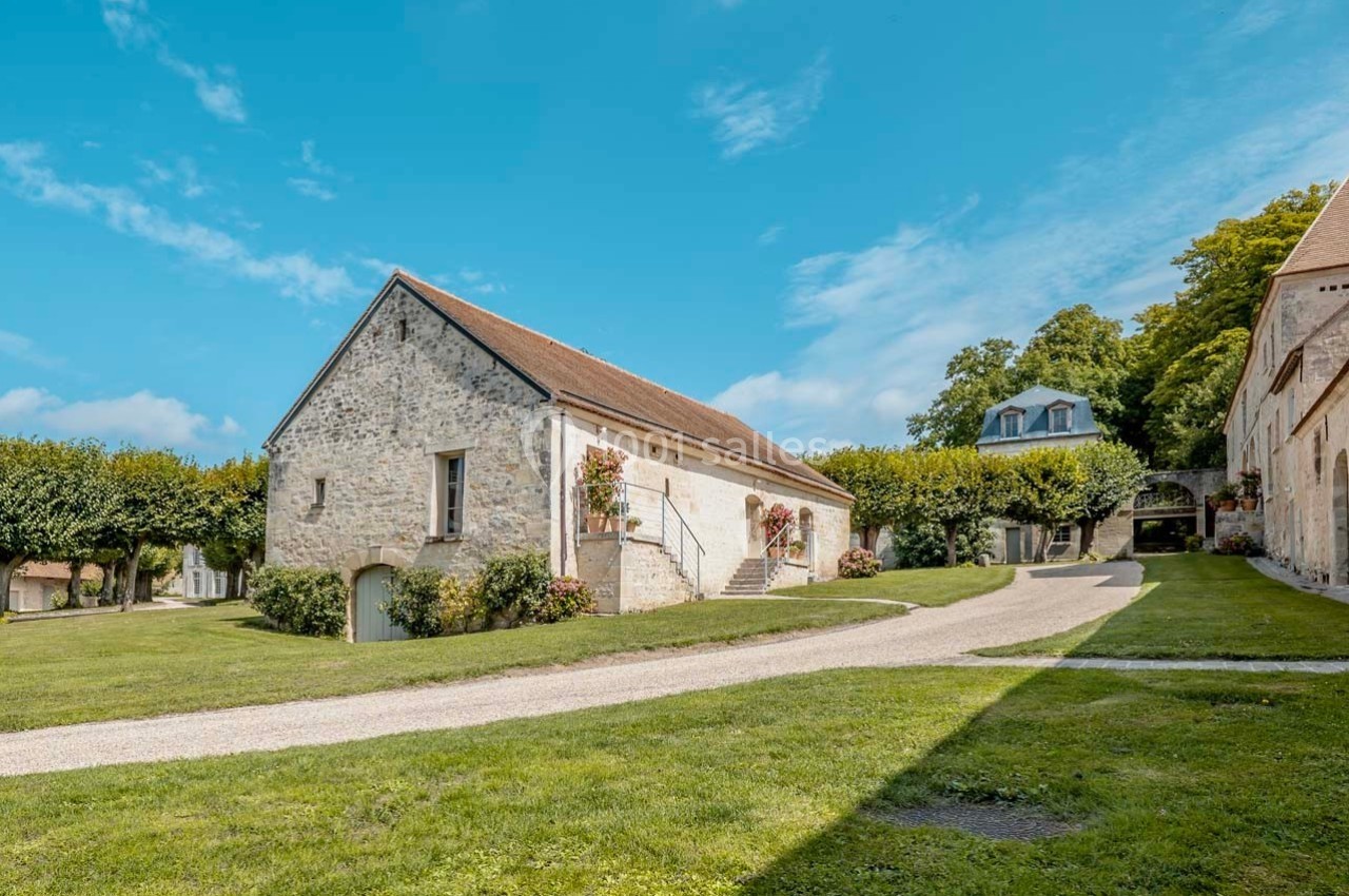 Bâtiment en pierre avec toit en pente, entouré de pelouses et d'arbres, sous un ciel bleu dégagé.