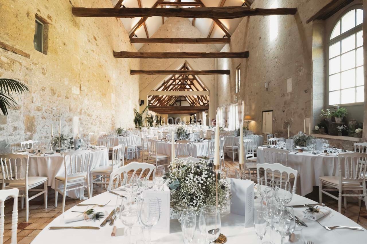 Salle de réception lumineuse avec tables rondes décorées de fleurs blanches, chaises blanches et poutres apparentes.