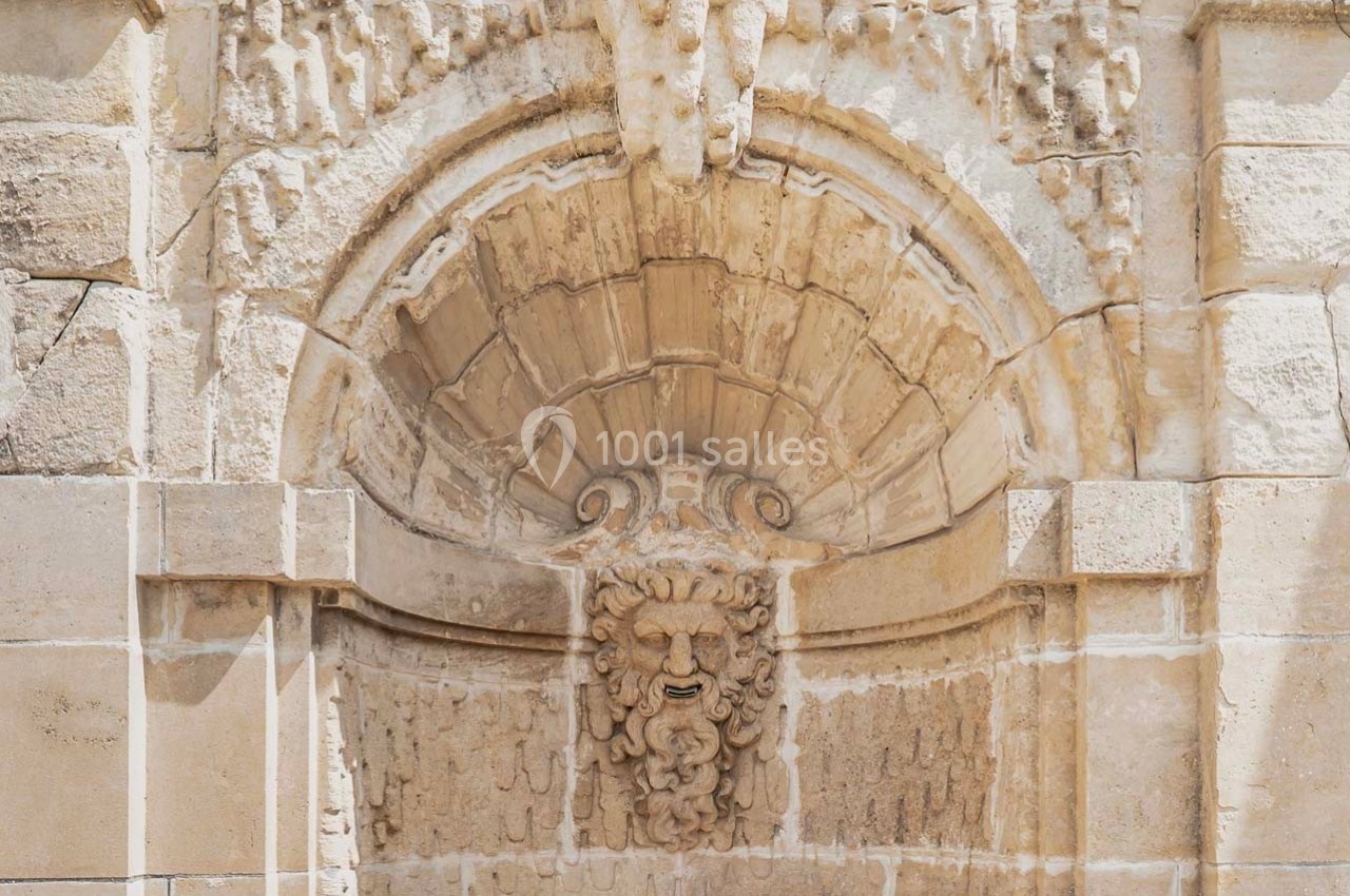 Bas-relief en pierre représentant un visage sculpté sous une arche ornée, intégré à une façade ancienne.