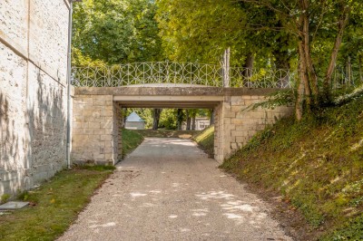 Petit pont en pierre surplombant un chemin, entouré de verdure et jouxtant un bâtiment ancien avec toiture en ardoise.