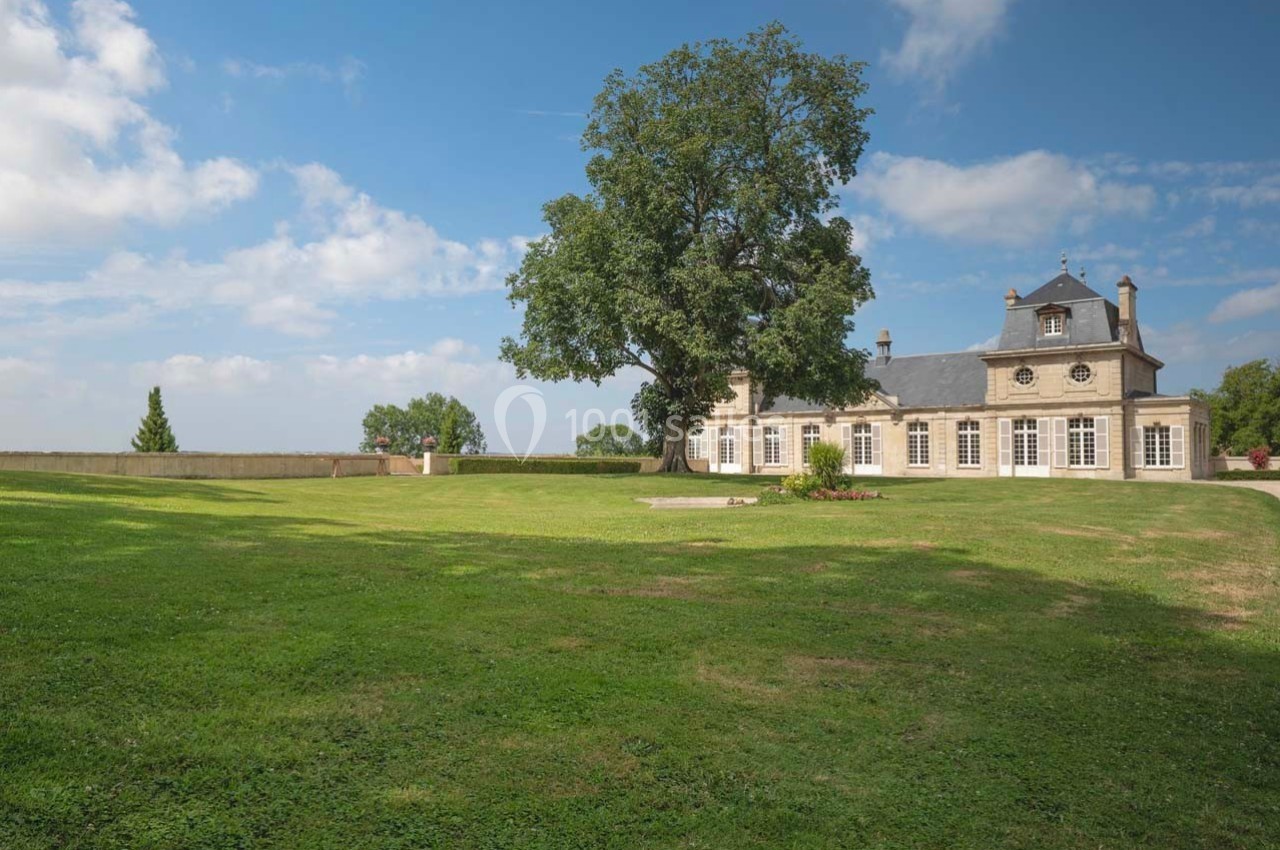 Grand jardin verdoyant avec un grand arbre au centre, devant une maison en pierre sous un ciel partiellement nuageux.