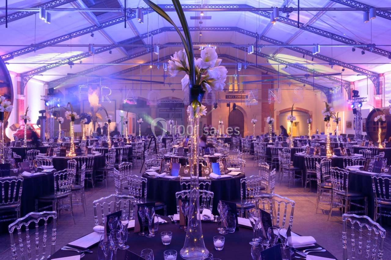 Salle de réception décorée avec des tables rondes, des chaises transparentes et des arrangements floraux sous un éclairage…