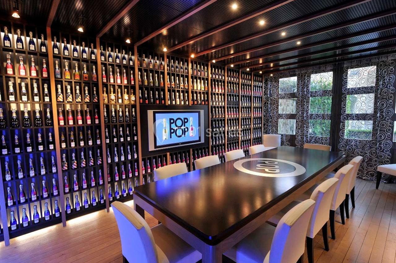 Salle de réunion moderne avec une grande table entourée de chaises, des étagères remplies de bouteilles et un écran…