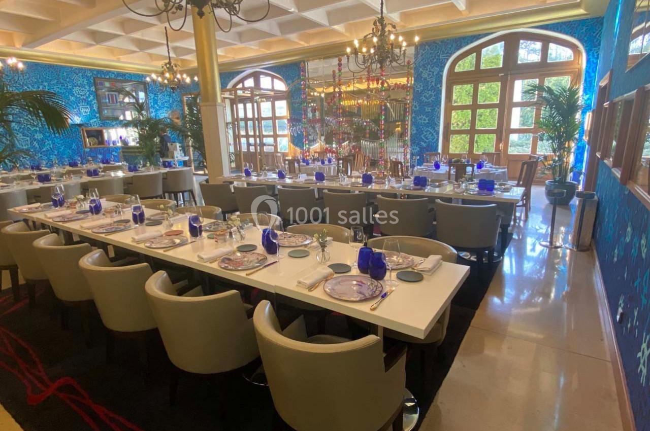Salle de restaurant élégante avec tables dressées, chaises beige, murs bleus décorés et grandes fenêtres lumineuses.