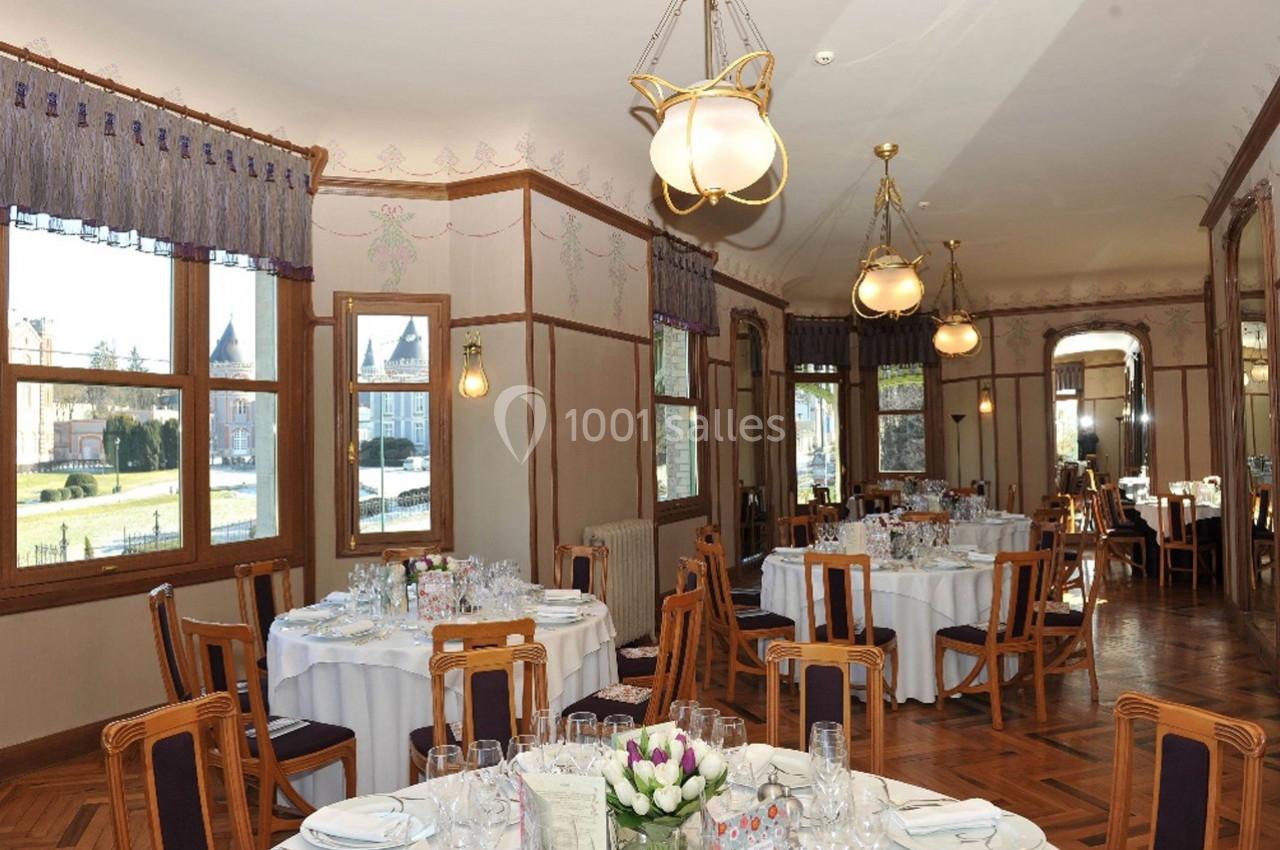 Salle de restaurant élégante avec tables rondes dressées, grandes fenêtres et vue sur un jardin extérieur.