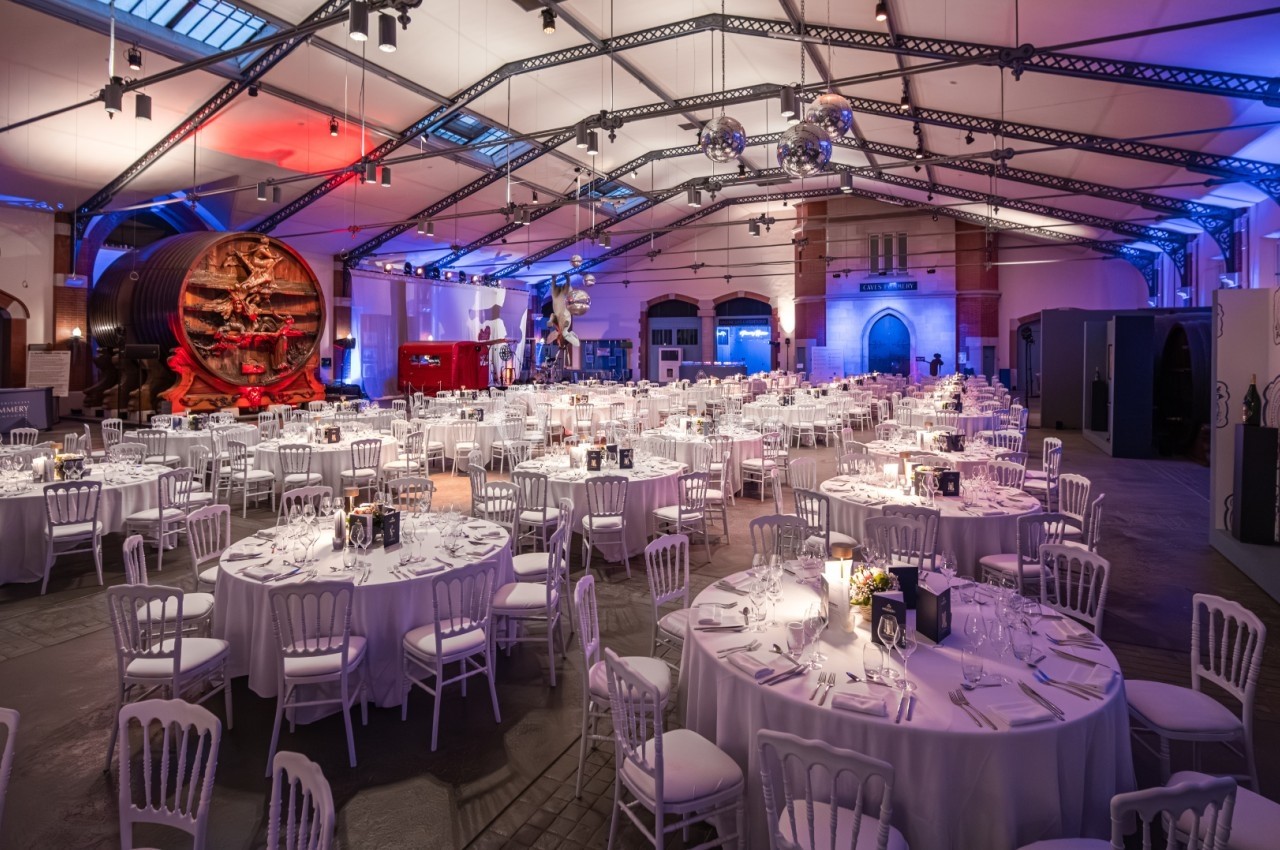 Salle de réception élégante avec tables rondes dressées, éclairage coloré et grand tonneau en arrière-plan.
