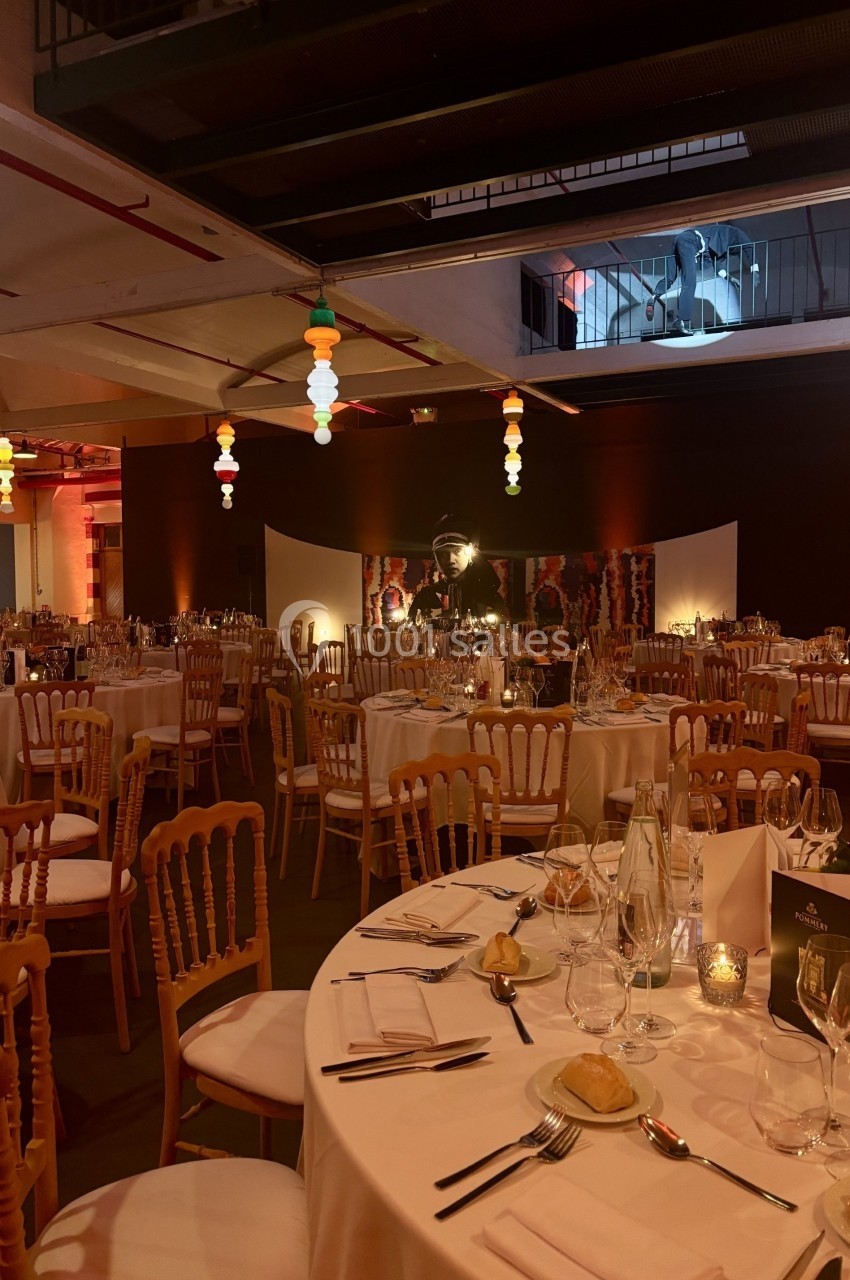 Salle de réception décorée avec des tables rondes dressées, chaises en bois et éclairage chaleureux.
