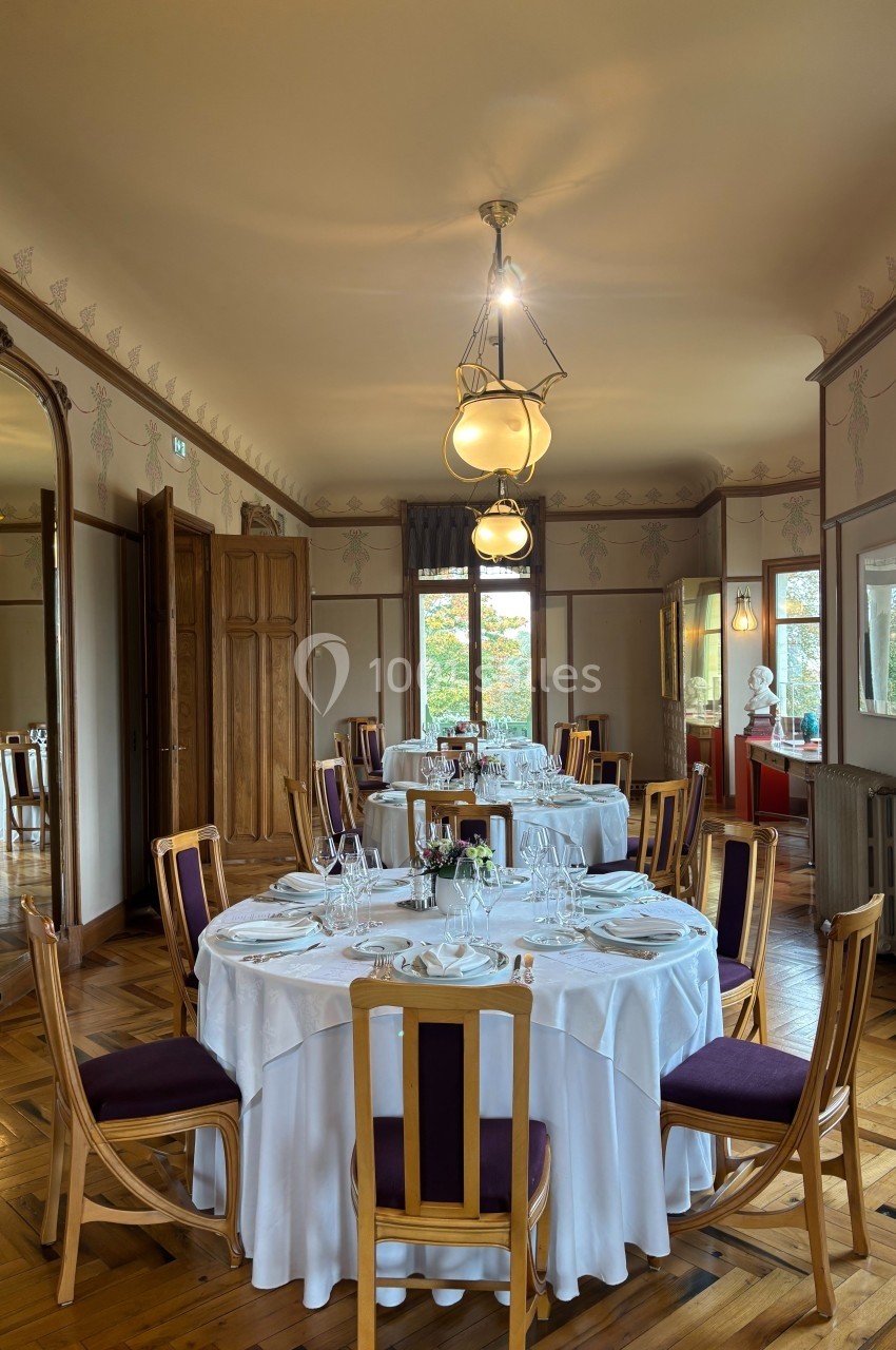 Salle de restaurant élégante avec tables rondes dressées, chaises en bois et lumière naturelle entrant par les fenêtres.