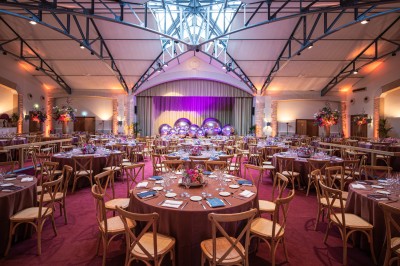 Salle voûtée décorée avec des tables rondes dressées pour un événement, éclairée par des lumières violettes.