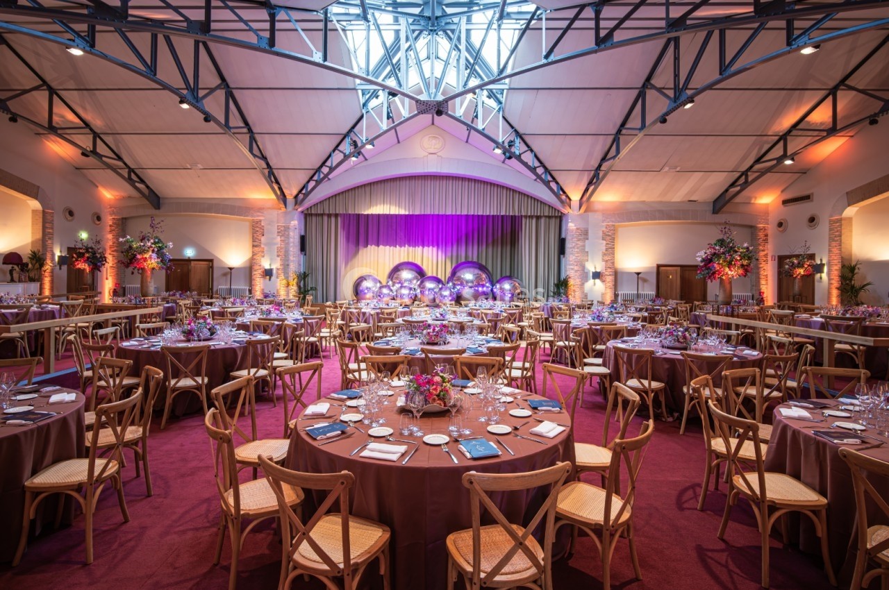 Salle de réception décorée avec des tables rondes, chaises en bois, éclairage chaleureux et scène en arrière-plan.