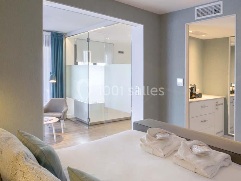 Chambre d'hôtel moderne avec lit double, salle de bain vitrée, fauteuil, table basse et machine à café.