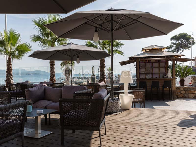 Terrasse en bois avec mobilier d'extérieur, parasols, palmiers et vue sur un port maritime en arrière-plan.