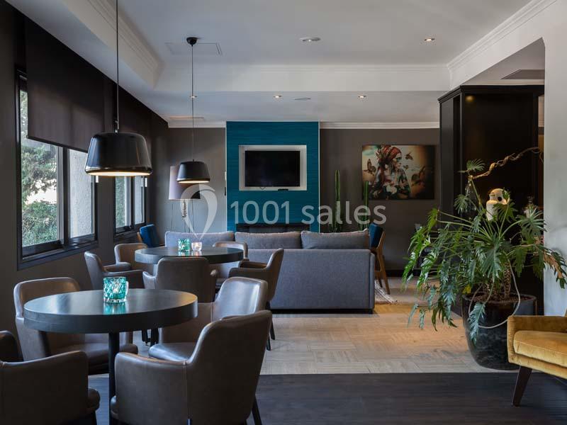 Salle moderne avec tables, chaises en cuir, canapé, plantes, éclairage suspendu et télévision murale.