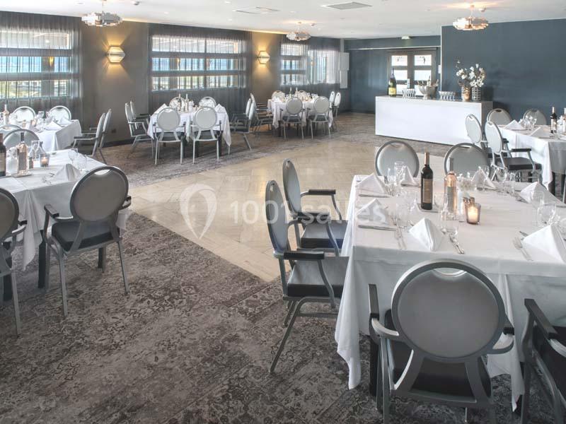 Salle de restaurant élégante avec tables dressées, nappes blanches, chaises grises et lumière naturelle entrant par les…