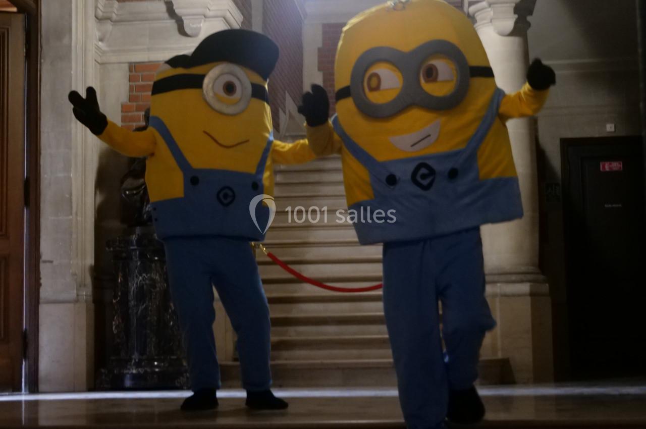 Deux personnes déguisées en personnages jaunes avec salopettes bleues dans un bâtiment avec un escalier en arrière-plan.