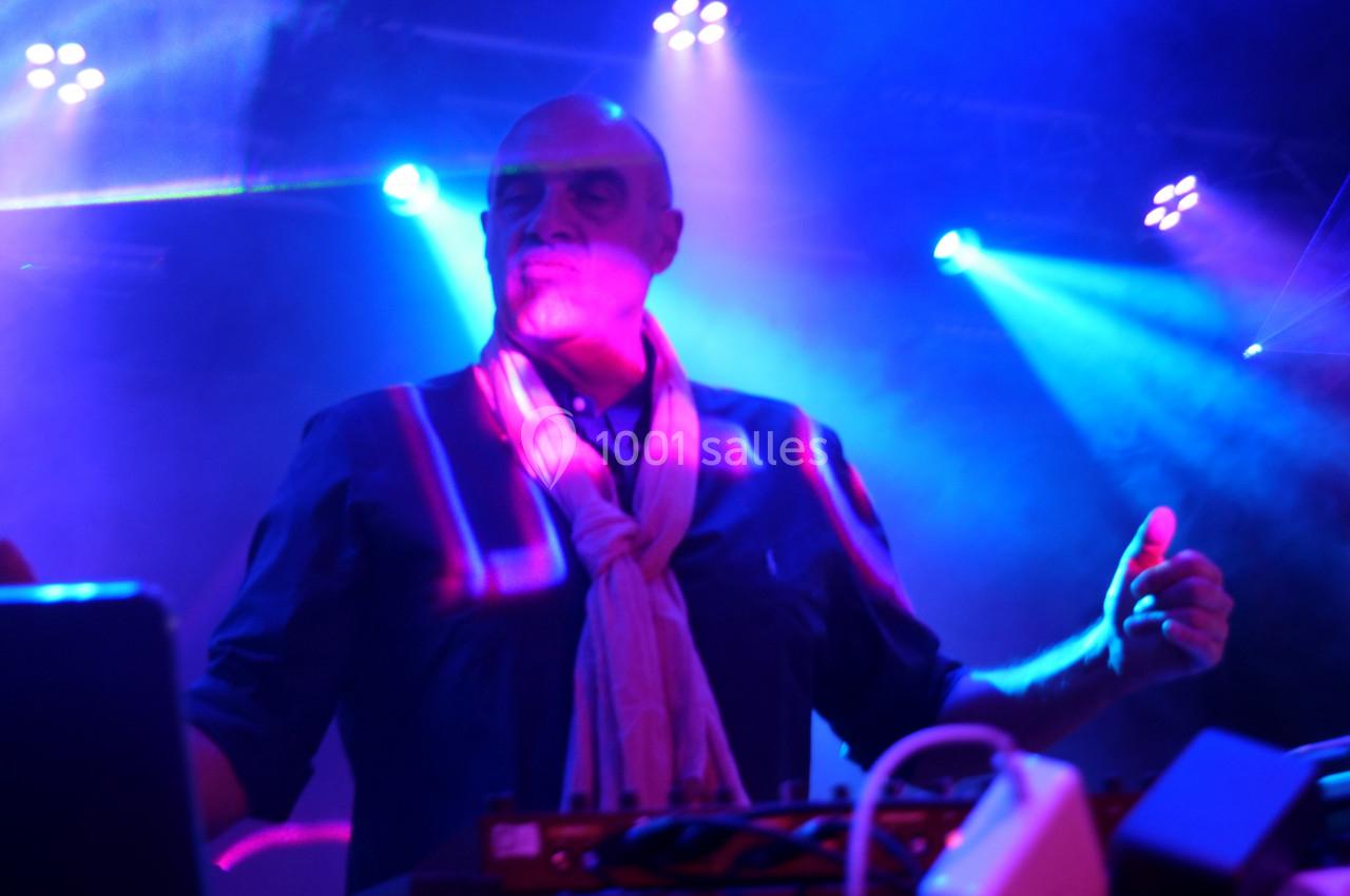 Homme debout sur scène, éclairé par des lumières bleues et roses, manipulant un équipement électronique.