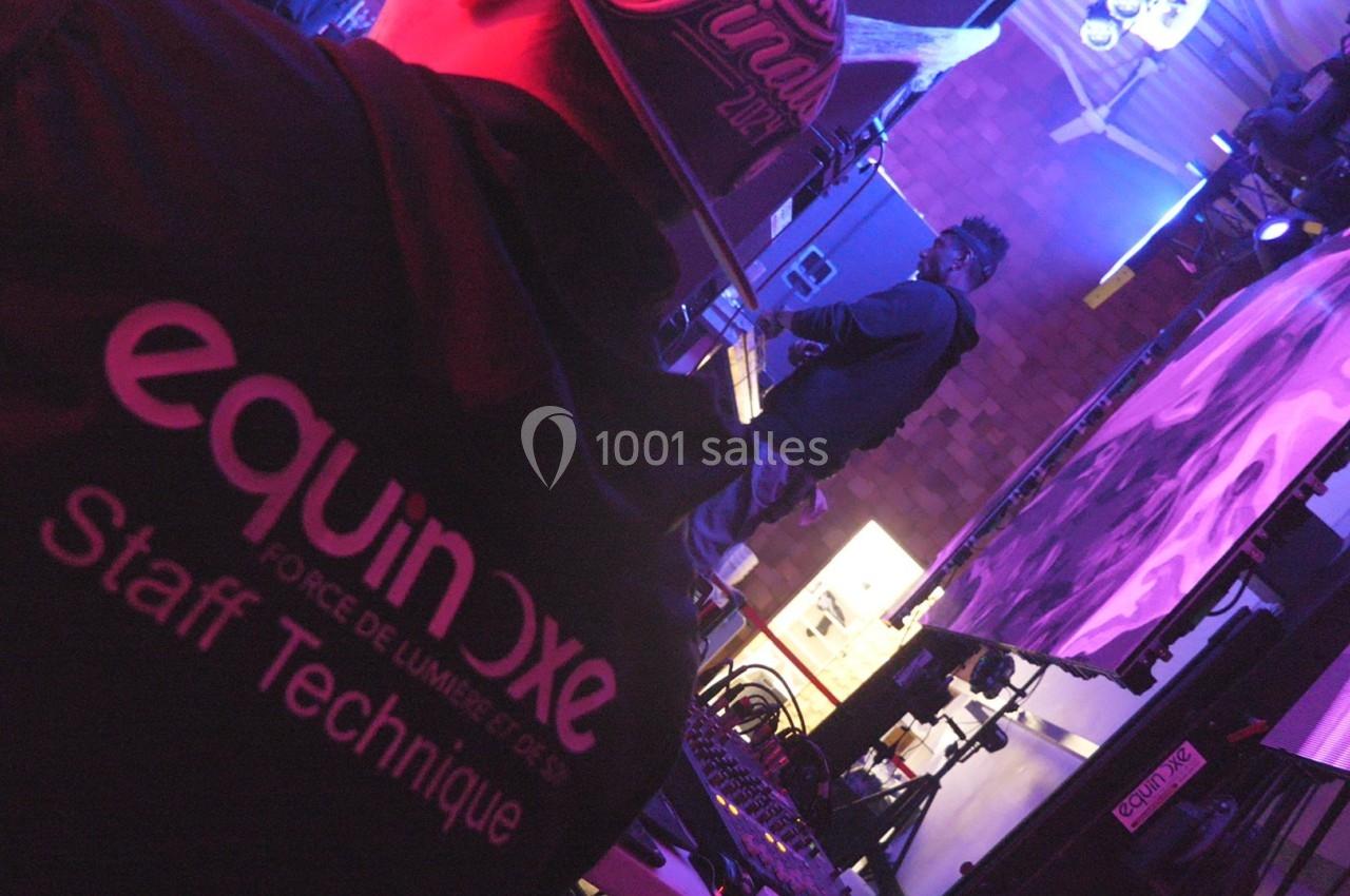 Technicien en veste ’Staff Technique’ supervisant une scène avec un musicien et des éclairages colorés en arrière-plan.