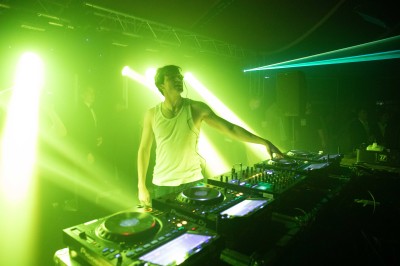 Un DJ mixe sur une scène éclairée, avec un écran lumineux affichant des motifs rouges en arrière-plan.