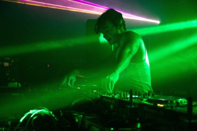 Un DJ mixe sur une scène éclairée, avec un écran lumineux affichant des motifs rouges en arrière-plan.