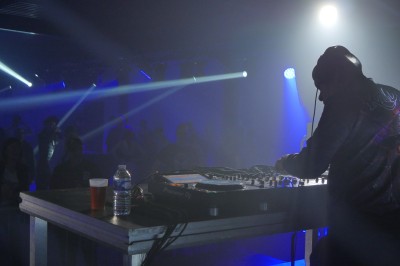 Un DJ mixe sur une scène éclairée, avec un écran lumineux affichant des motifs rouges en arrière-plan.