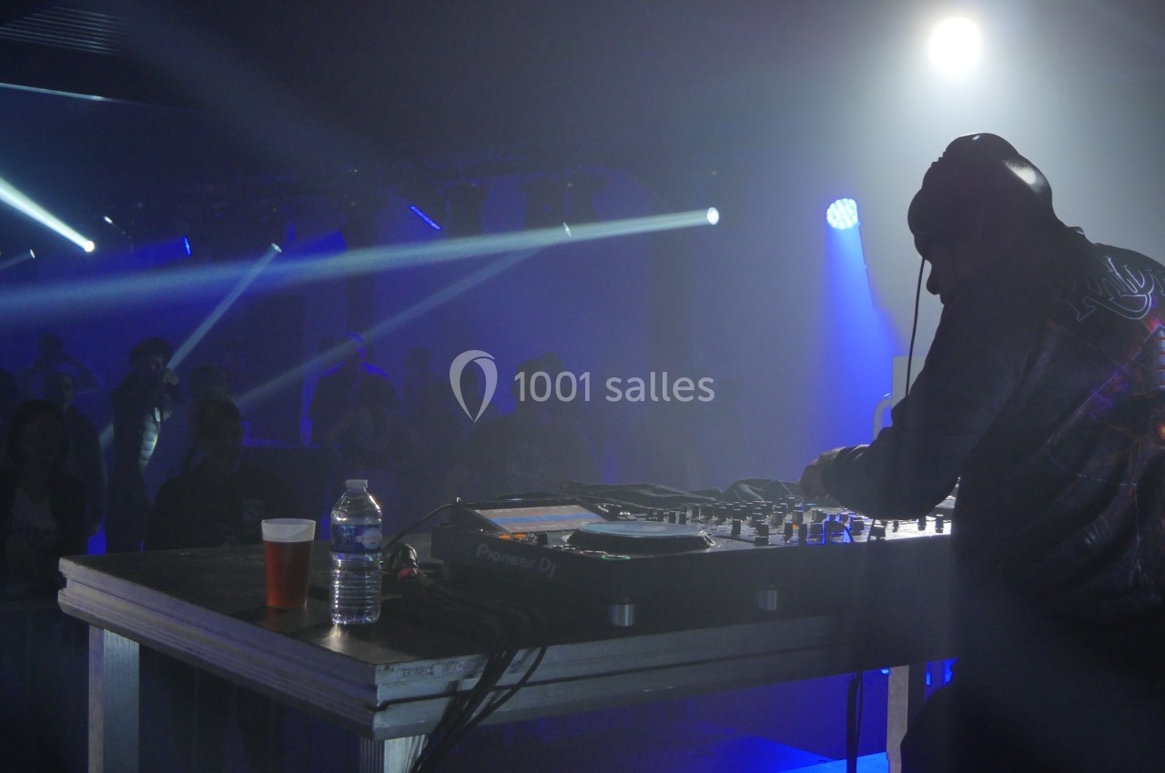 Un DJ mixe sur scène dans une ambiance sombre avec des éclairages bleus et un public en arrière-plan.