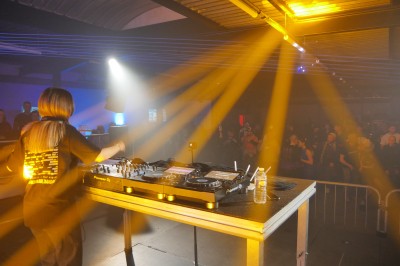 Un DJ mixe sur une scène éclairée, avec un écran lumineux affichant des motifs rouges en arrière-plan.