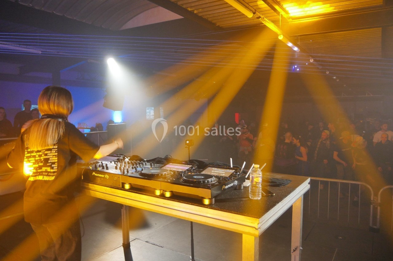 Une DJ mixe sur une scène éclairée par des projecteurs jaunes, face à un public dans une salle sombre.