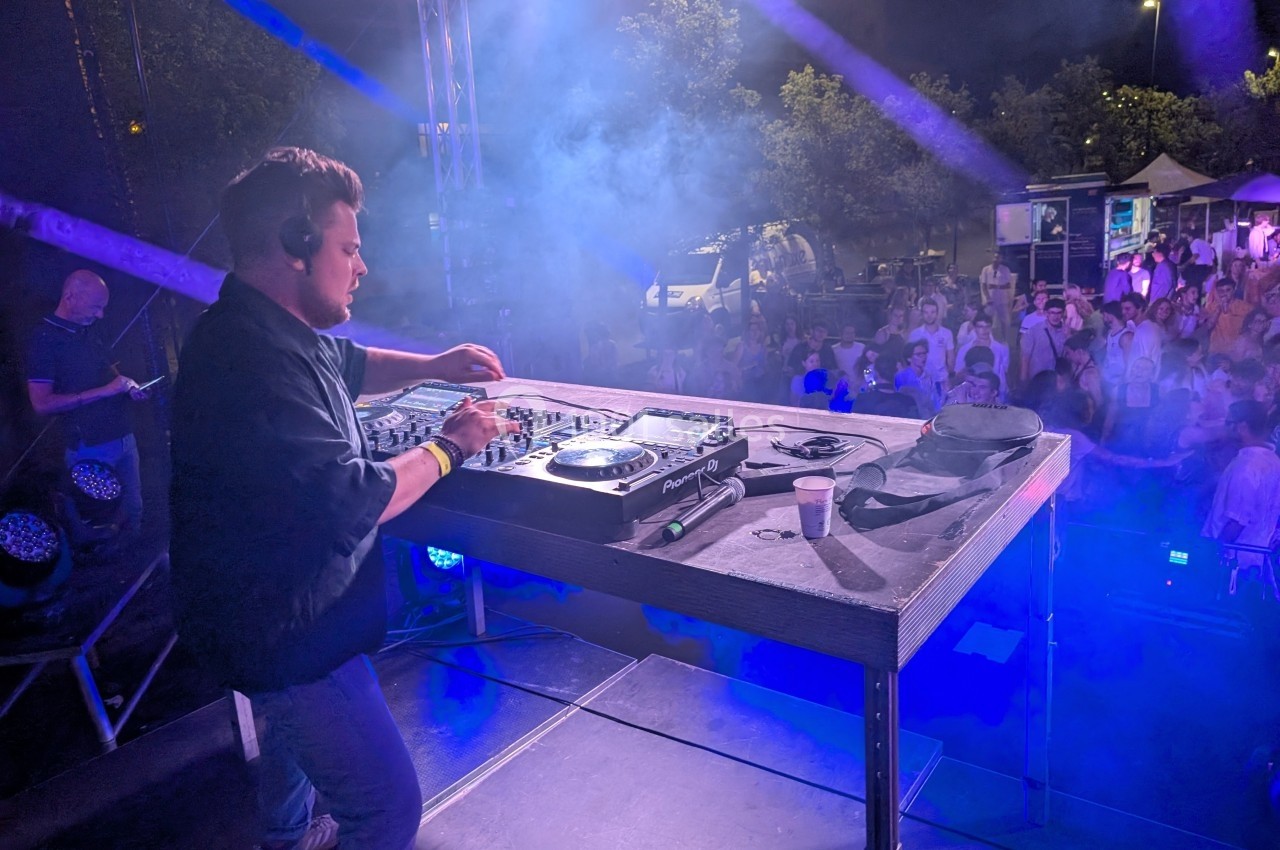 Un DJ mixe sur une scène en plein air, face à une foule sous des lumières colorées, de nuit.