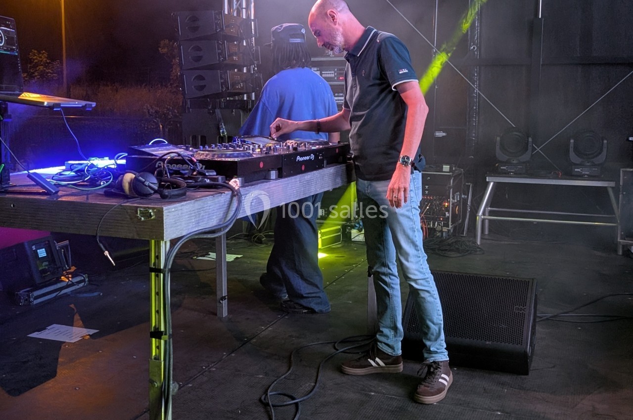 Un DJ ajuste les commandes d'une table de mixage sur une scène extérieure éclairée par des projecteurs colorés.