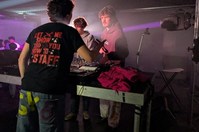 Un homme en t-shirt noir échange avec deux autres personnes près d'une table de mixage dans une salle éclairée par des…