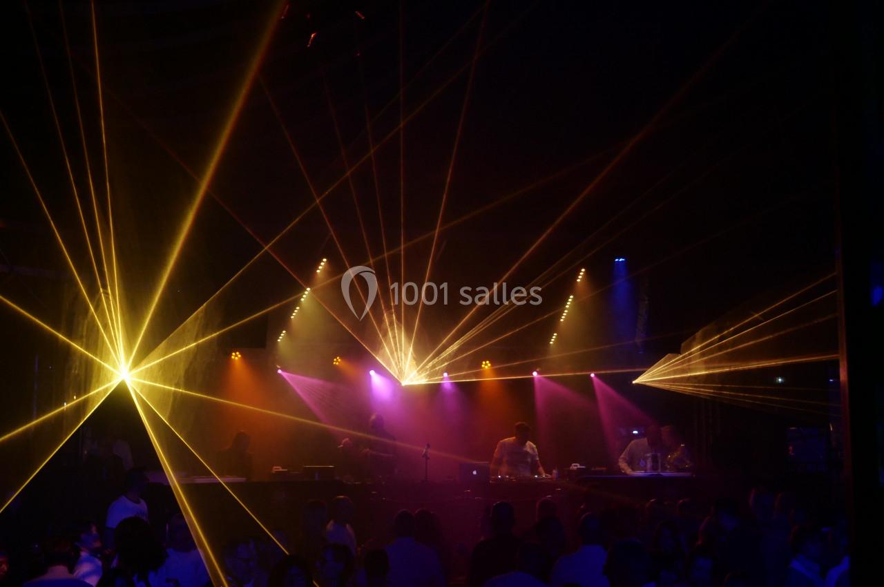 Salle sombre animée avec des faisceaux lumineux colorés et des personnes dansant devant une scène avec des DJ.