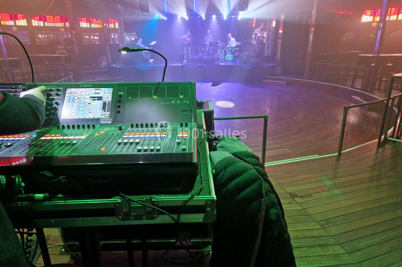 Console de mixage audio en premier plan, avec une scène éclairée et un groupe en répétition en arrière-plan.