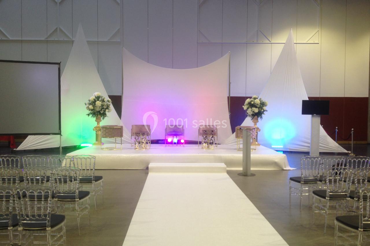 Scène décorée pour un événement avec chaises transparentes, fleurs, éclairages colorés et fond en tissu blanc.