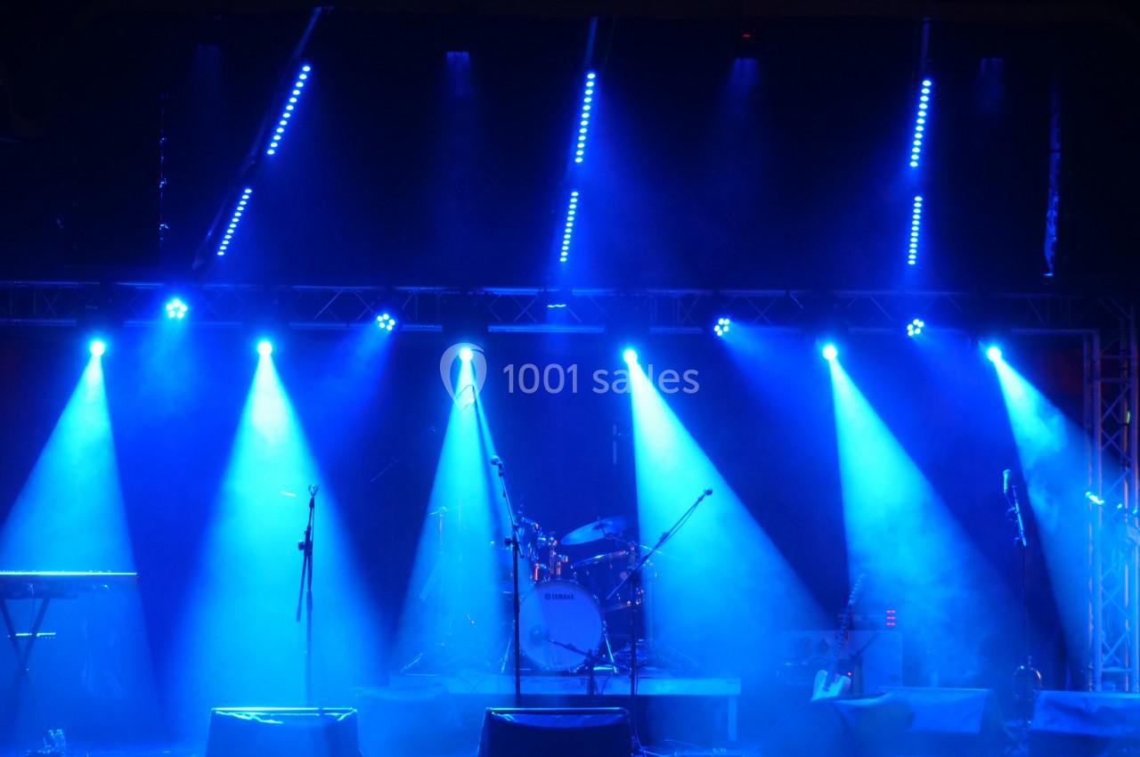 Scène de concert vide éclairée par des projecteurs bleus, avec une batterie et des micros installés.