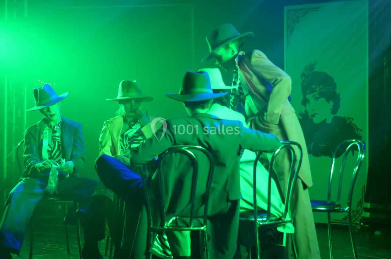 Quatre personnes en costumes et chapeaux assises sur des chaises, éclairées par une lumière verte dans une ambiance scénique.
