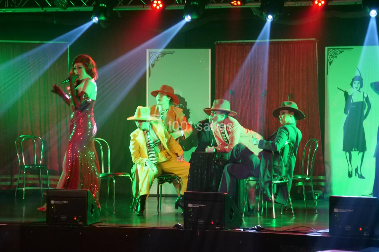 Une chanteuse en robe rouge performe sur scène devant des hommes en costumes assis, éclairés par des projecteurs colorés.