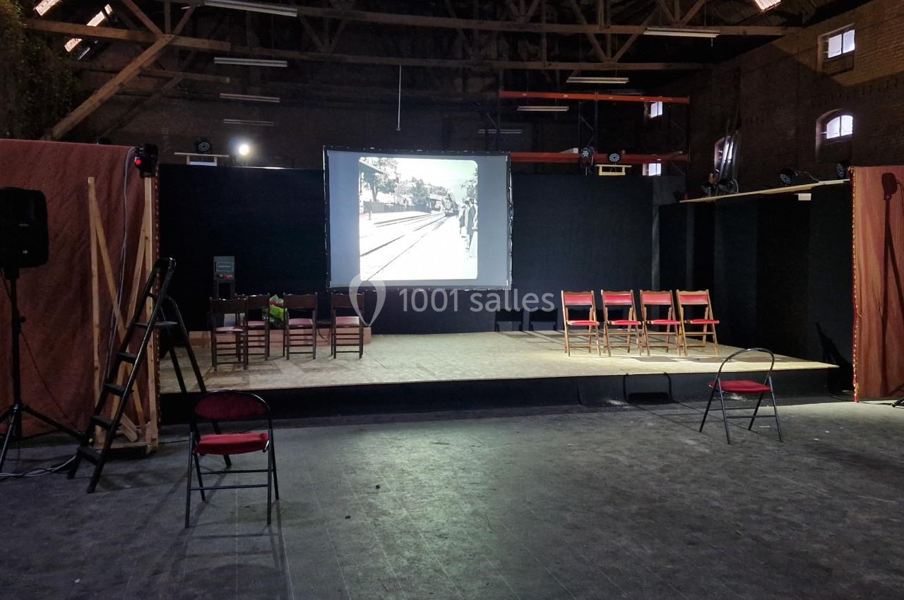 Salle sombre avec une scène équipée d'un écran de projection, des chaises pliantes et un éclairage tamisé.