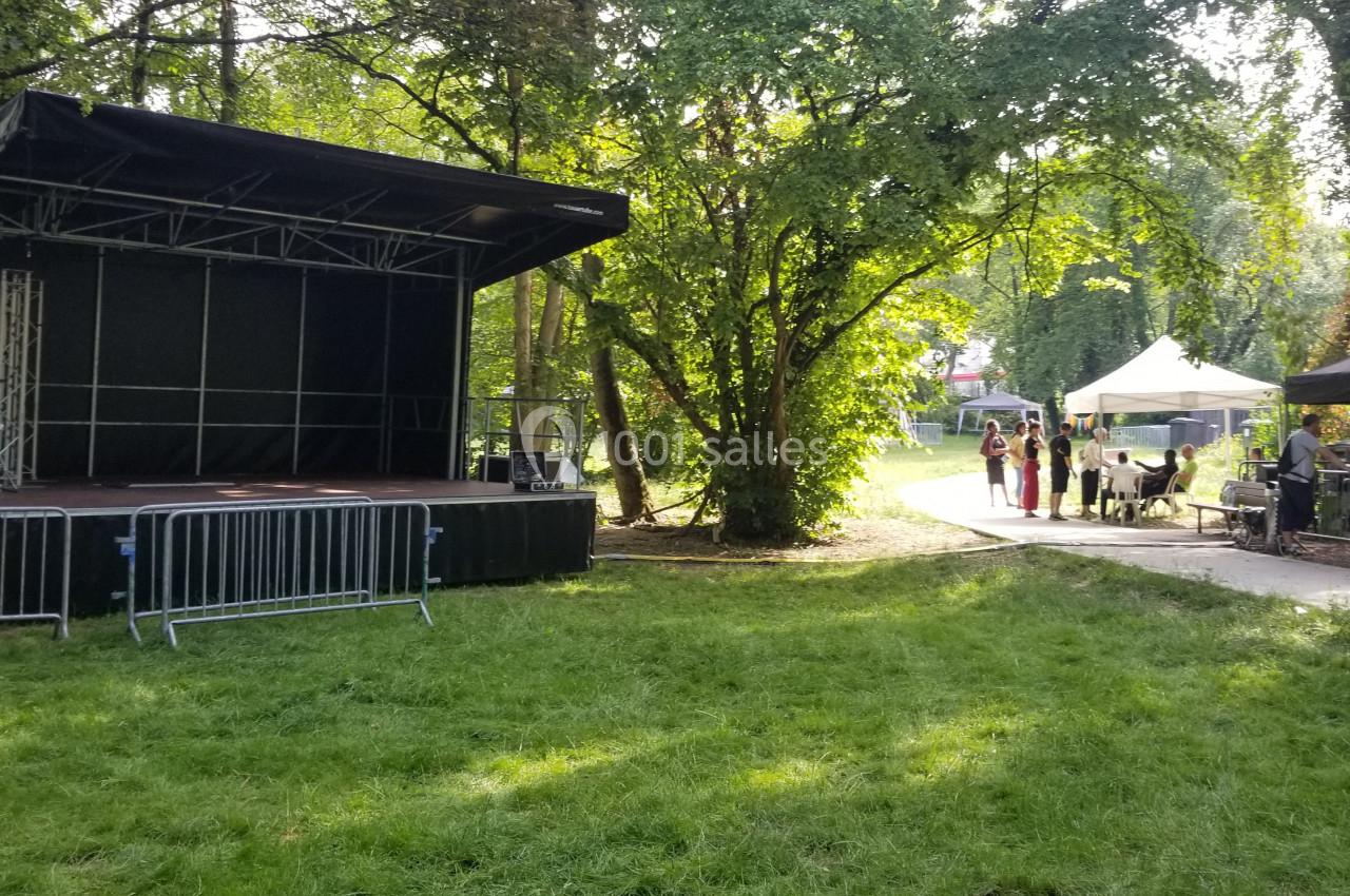 Scène extérieure vide entourée d'arbres, avec des tentes et des personnes en arrière-plan sur un chemin.