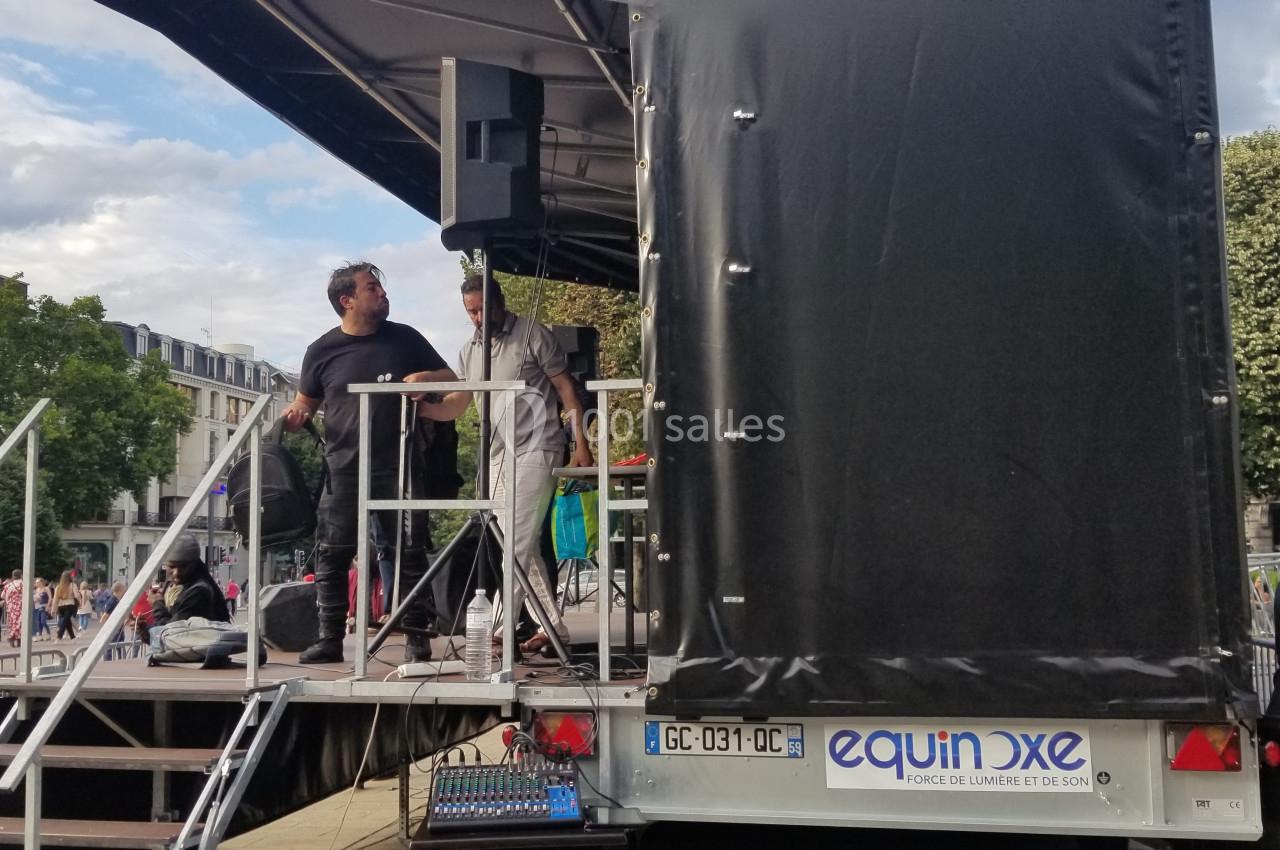 Deux hommes sur une scène mobile en plein air, avec du matériel audio, entourés de spectateurs et d'arbres.