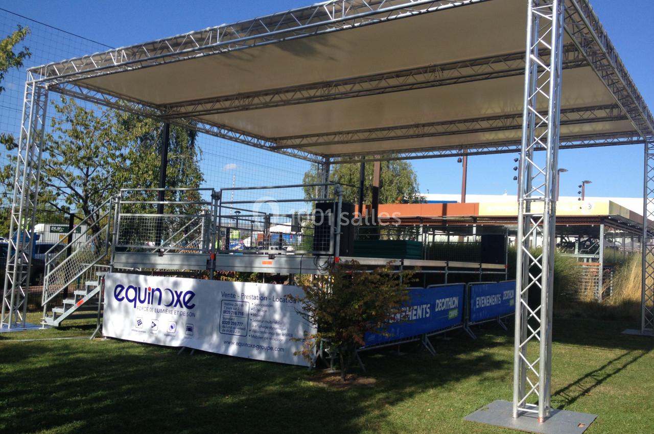 Structure métallique avec toit en toile, installée sur une pelouse, entourée de barrières et de panneaux publicitaires.