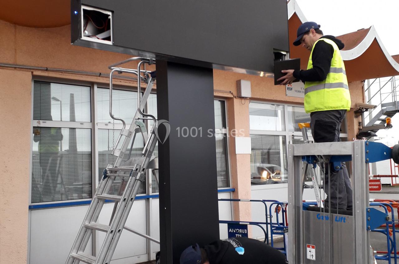 Deux techniciens installent un panneau numérique sur un support métallique devant un bâtiment.