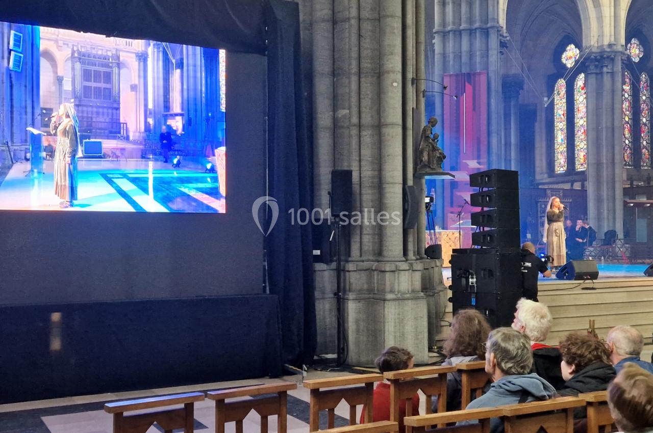 Un écran diffuse une performance artistique dans une église, devant un public assis sur des bancs en bois.