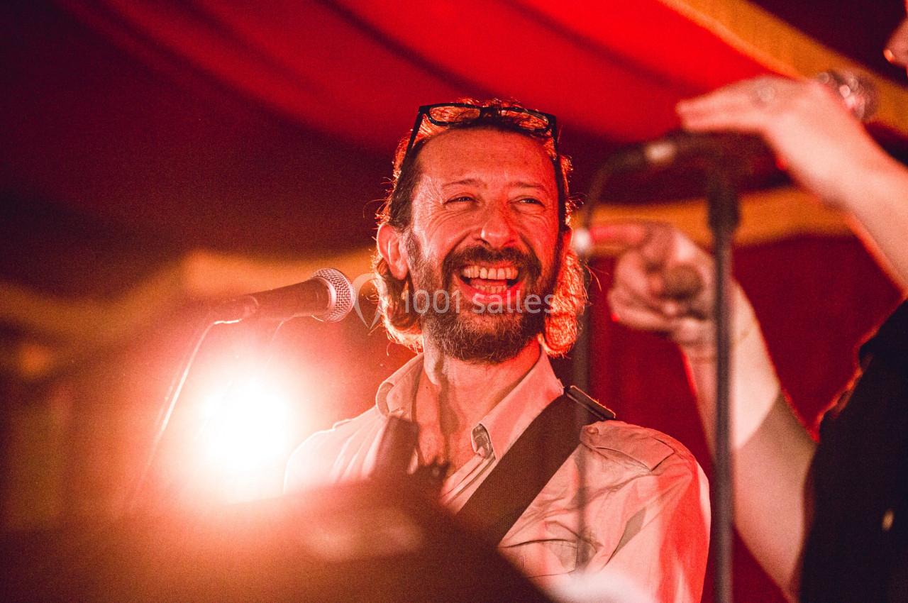 Un homme souriant chante ou parle au micro sous un éclairage chaleureux, avec un rideau rouge en arrière-plan.