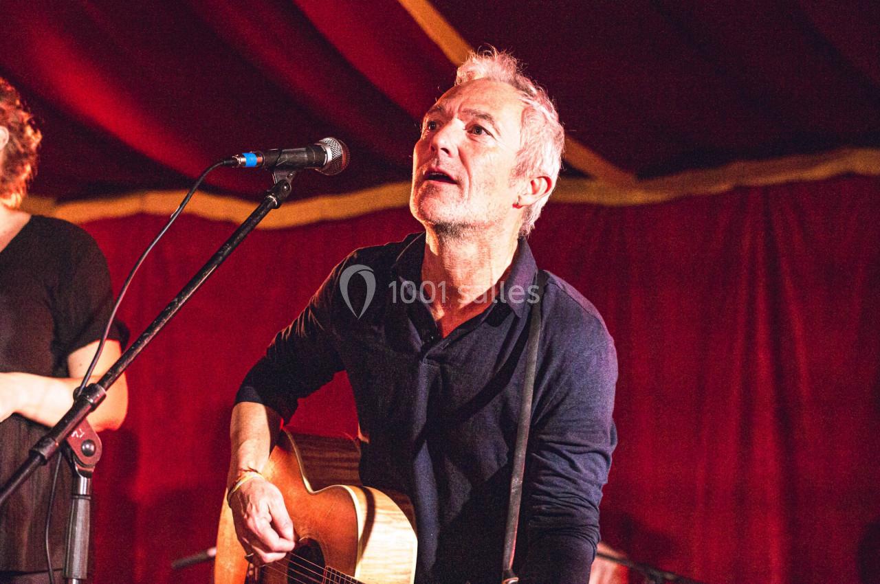 Un homme joue de la guitare acoustique sur scène, devant un rideau rouge, avec un micro à proximité.