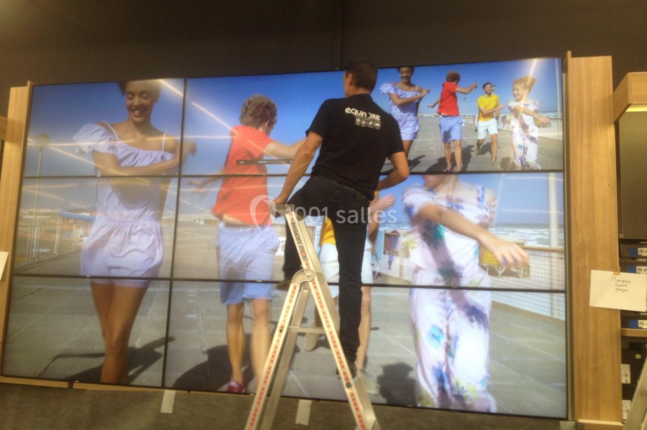 Un technicien ajuste un mur d'écrans affichant des personnes marchant sur une jetée en bord de mer.