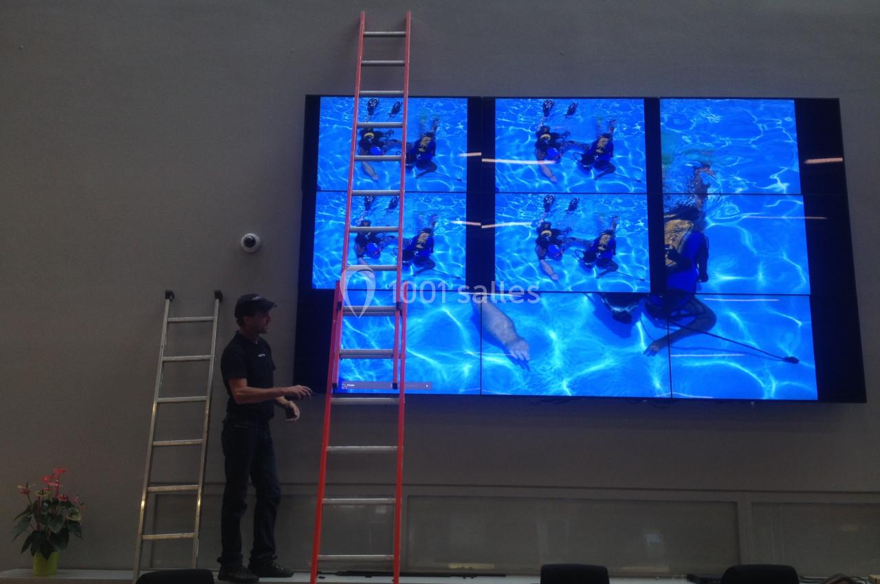 Un technicien ajuste un mur d'écrans affichant des images sous-marines, avec deux échelles à proximité.