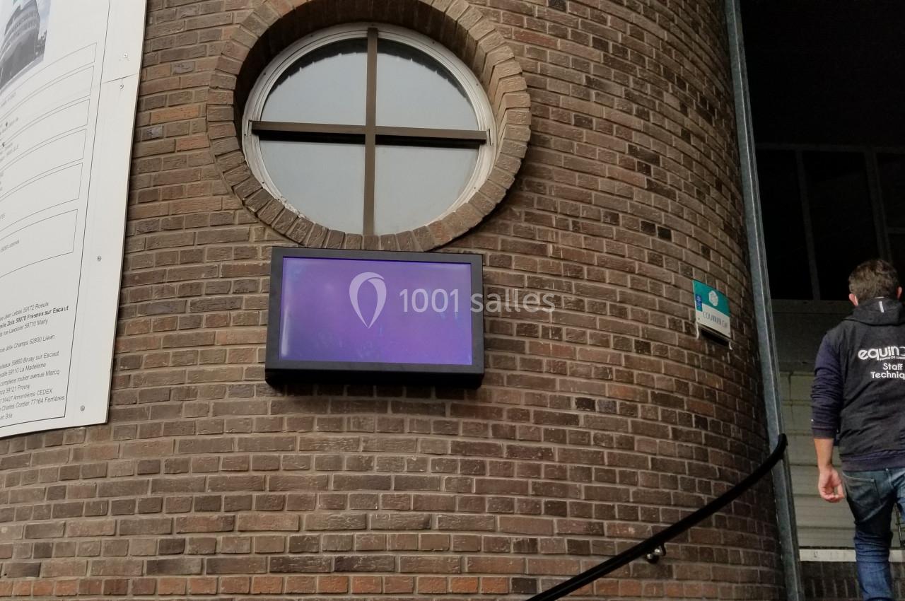 Écran numérique fixé sur un mur en briques près d'une porte, avec une personne montant des marches adjacentes.