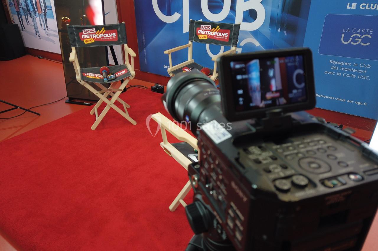 Une caméra filme deux fauteuils sur un tapis rouge dans un espace promotionnel avec des affiches en arrière-plan.