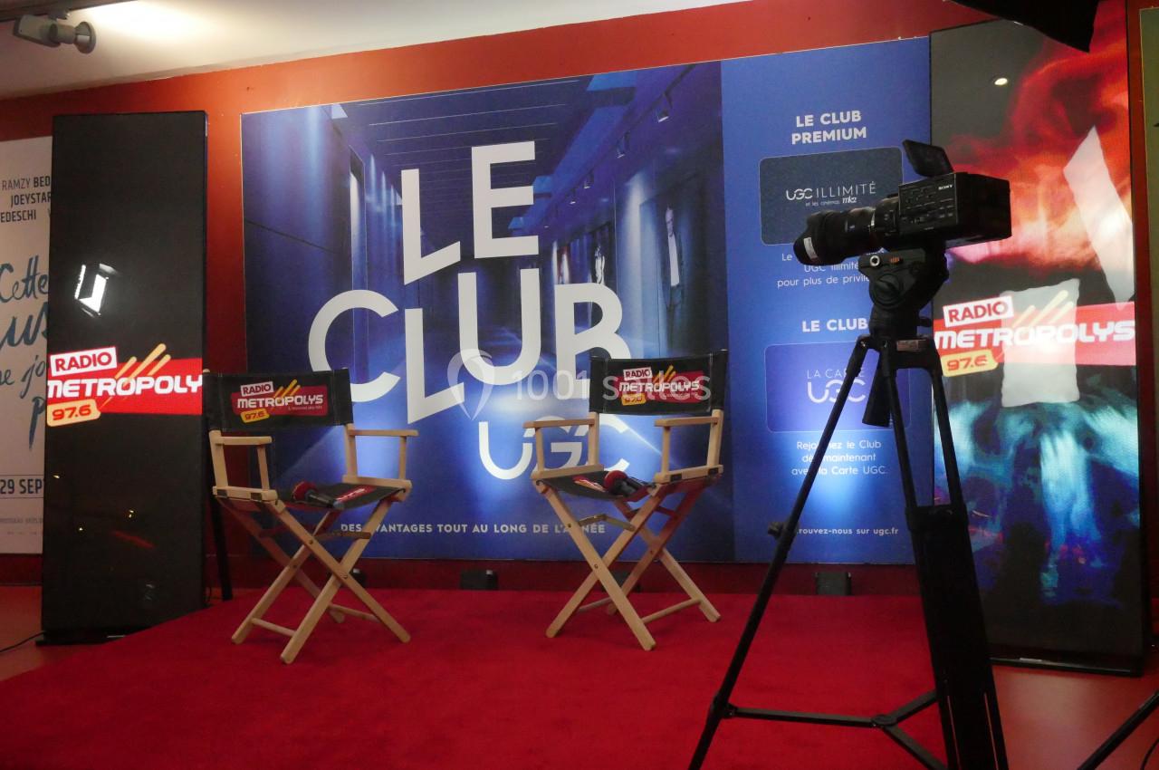 Deux chaises sur un tapis rouge devant un fond promotionnel ’Le Club UGC’, avec une caméra et des logos de radio.